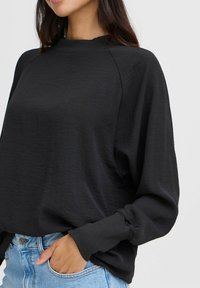 Maglietta nera a maniche lunghe con scollatura rotonda a coste e texture morbida. Presenta polsini elastici e una vestibilità comoda, abbinata a jeans in denim azzurro chiaro.