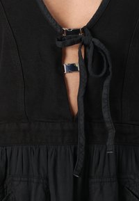 Abito nero con scollo a V profondo, chiusura con nastro di stoffa e accenti metallici. Parte superiore testurizzata con fondo arricciato e leggero lucido.