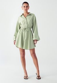 Šviesiai žalia mygtukinė suknelė su ilgomis rankovėmis, elastinga juosmens linija ir plačiu sijonu. Pagaminta iš lygaus audinio su laisvu siluetu.