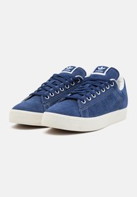 Sapatilhas azuis de camurça com sola de borracha branca, apresentando detalhes perfurados nas laterais e atacadores azuis a condizer. Logótipo da Adidas na lingueta.