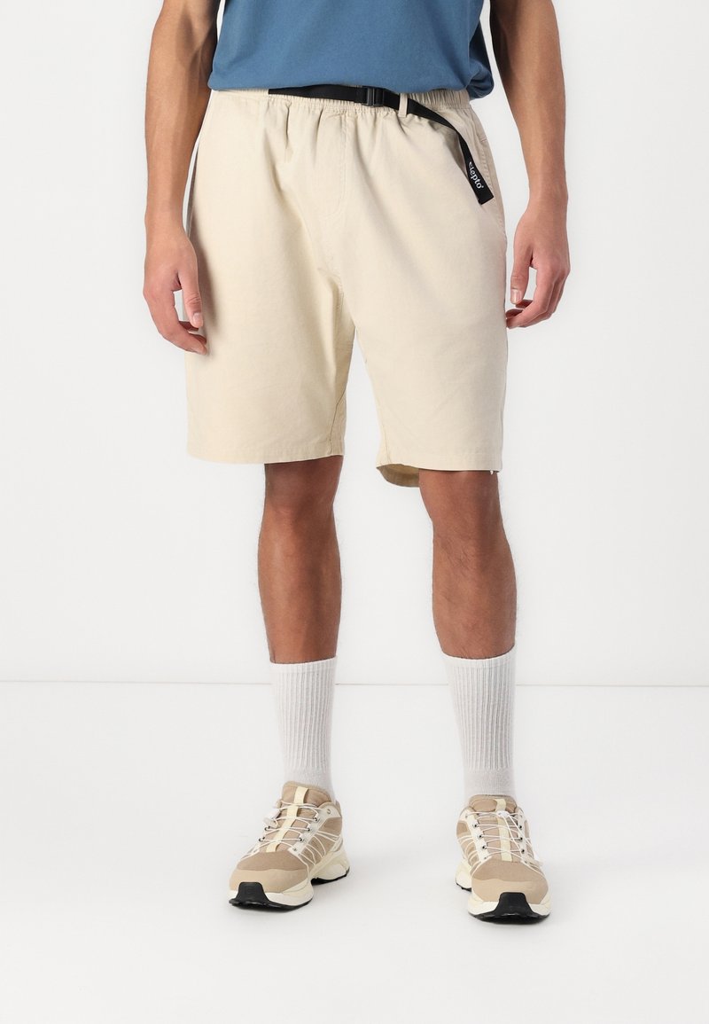 Homme portant un short beige au genou avec une ceinture noire, des chaussettes blanches, des baskets beiges et une chemise bleue, debout devant un fond blanc uni.