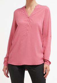 Femme portant un chemisier rose pâle à manches longues avec un col en V et une patte de boutonnage, associé à un pantalon noir sur un fond blanc.