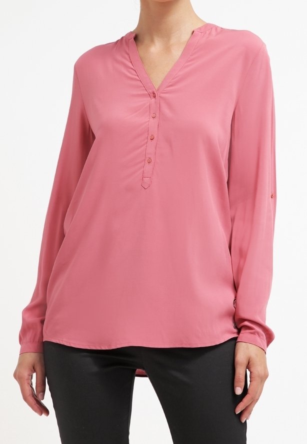 Femme portant un chemisier rose pâle à manches longues avec un col en V et une patte de boutonnage, associé à un pantalon noir sur un fond blanc.