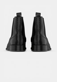 O pereche de ghete Chelsea negre din piele cu detalii brogue, prezentate din spate pe un fundal alb.