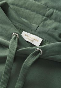 Gros plan sur le tissu d'un sweat à capuche vert montrant des cordons épais avec œillets métalliques et une étiquette blanche indiquant « JACK & JONES Originals » à l'intérieur du col.