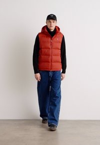 Rød puffervest med hette, med glidelås foran og logo detaljer, brukt over en svart langermet skjorte og kombinert med baggy blå jeans.