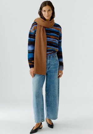 Pull en maille à rayures bleues, brunes et oranges ; associé à un jean ample bleu clair et une écharpe en maille brune. Ballerines noires à bouts pointus.