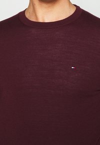 Bordeauxroter Strickpullover mit rundem Ausschnitt, dezentem Muster und kleinem besticktem Logo in Rot, Weiß und Blau auf der Brust.