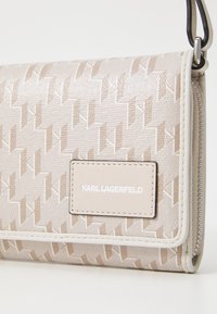 Beige wallet with a textured surface, featuring a geometric pattern, white leather accents, and a branded logo plaque. Zippered closure detail.