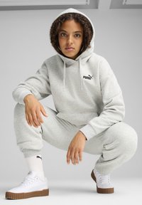 Joven con pelo rizado agachado vistiendo una sudadera ligera gris de Puma y pantalones de chándal, calcetines blancos de Puma y zapatillas blancas con suelas de goma.