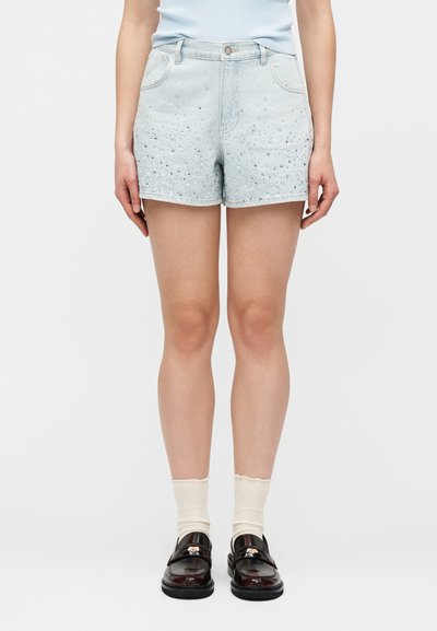 Γυναίκα που φοράει ανοιχτό μπλε τζιν σορτς με διακοσμητικά στρας, συνδυασμένα με λευκές κάλτσες και λαμπερά σκούρα καφέ μοκασίνια.