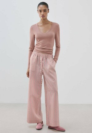 Mujer de pie con camiseta rosa de manga larga, pantalones de pierna ancha y cintura alta con rayas verticales rojas y blancas, y zapatos planos rosas, fondo liso.