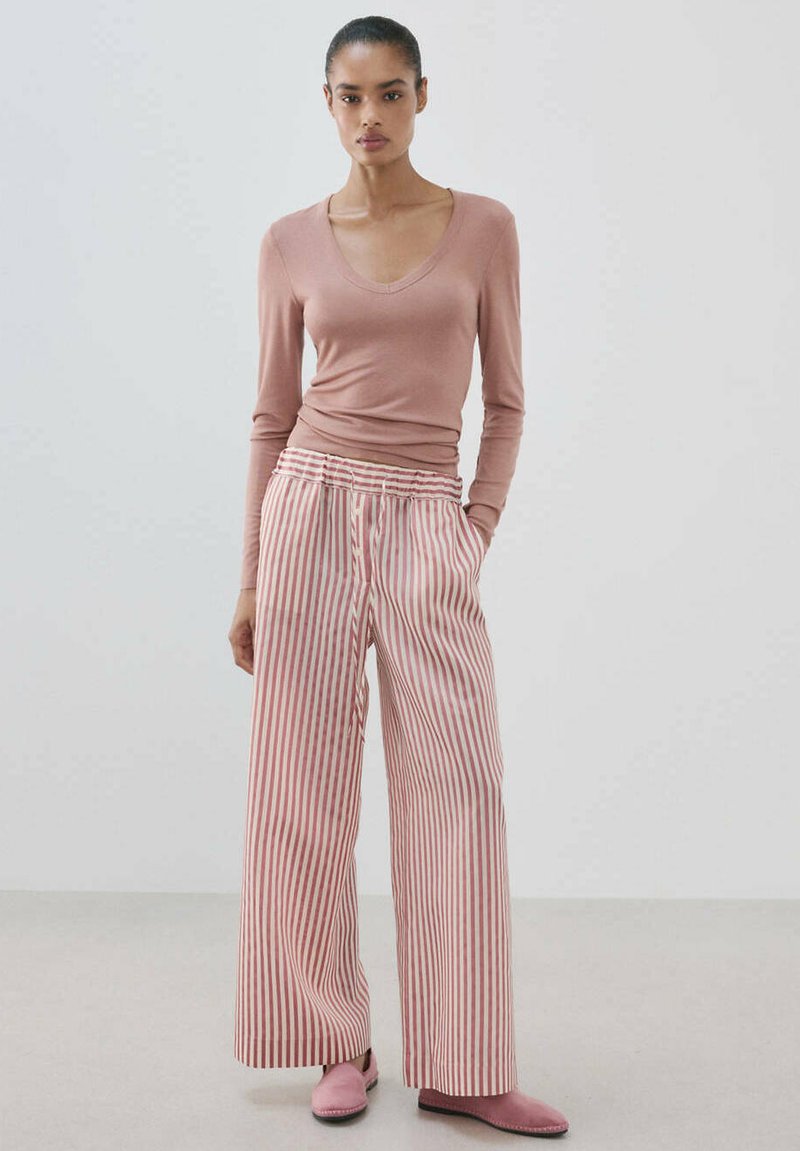Femme debout portant un haut rose à manches longues, pantalon large taille haute à rayures verticales rouges et blanches, et chaussures plates roses, arrière-plan uni.