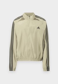 Béžová bunda na zips so stojatým golierom, čiernymi pruhmi na rukávoch a zošitým elastickým lemom. Na hrudi je logo Adidas.