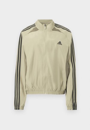 Beige zip-up jacka med ståkrage, svarta ränder på ärmarna och resår i fållen. Har en Adidas-logga på bröstet.