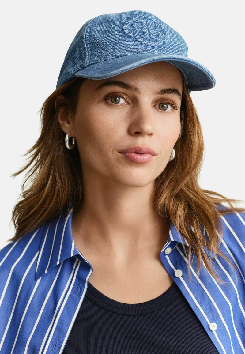 Femme aux cheveux châtain clair portant une casquette en jean bleue, des boucles d'oreilles argentées en forme d'anneau, une chemise rayée bleue et blanche sur un haut noir, expression neutre.