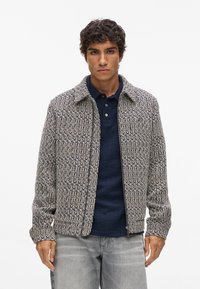 Textureret jakke med krave, intrikat vævet mønster i marineblå og beige, lynlås foran, båret over en mørk polo-shirt.