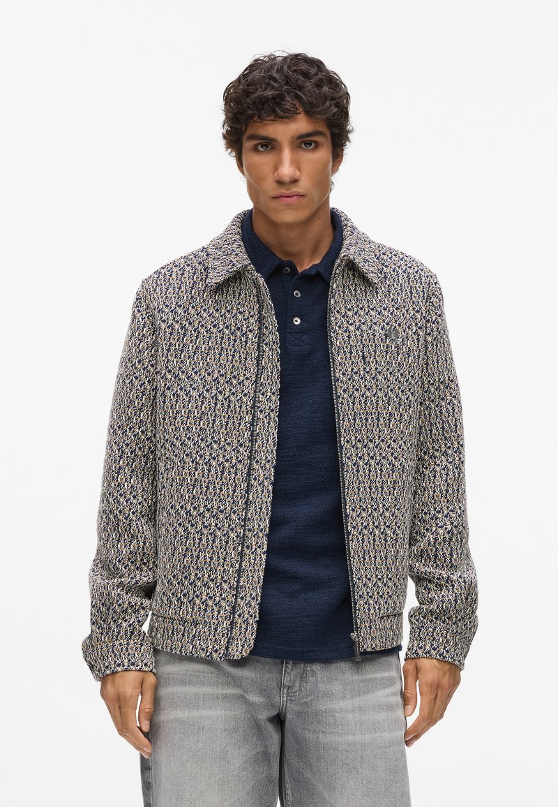 Textureret jakke med krave, intrikat vævet mønster i marineblå og beige, lynlås foran, båret over en mørk polo-shirt.