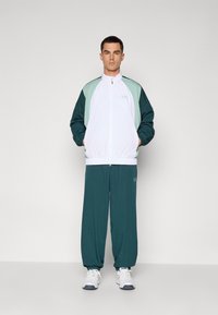 Ανδρική φόρμα που περιλαμβάνει ένα λευκό jacket με φερμουάρ και πράσινες-τιρκουάζ λεπτομέρειες, συνδυασμένο με τιρκουάζ παντελόνι. Κατασκευασμένο από ελαφρ�ύ ύφασμα, τα παπούτσια είναι λευκά.
