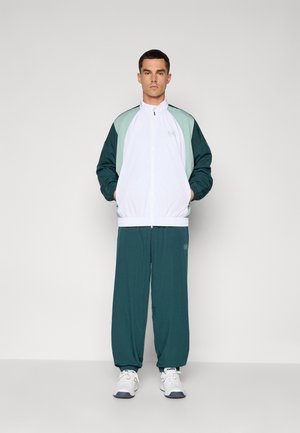 Training pentru bărbați, care include o jachetă albă cu fermoar, accentuată cu detalii verzi și teal, alături de pantaloni teal. Fabricat dintr-un material ușor, încălțămintea este albă.