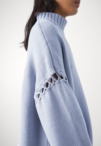 Personne portant un pull bleu clair en tricot avec un col montant et un motif ajouré sur le haut de la manche.