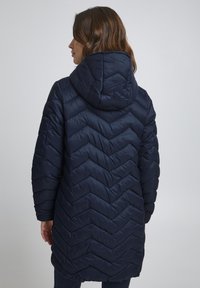Marinblå puffjacka med ett chevron-quiltat mönster, utrustad med huva och långa ärmar. Tyget verkar slätt och lätt.