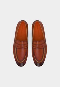 Ζευγάρι καφέ δερμάτινων μοκασίνων με ραφές και διακριτικές σκοτεινότερες αποχρώσεις στη μύτη και τη φτέρνα, απεικονισμένα από πάνω σε λευκό φόντο.