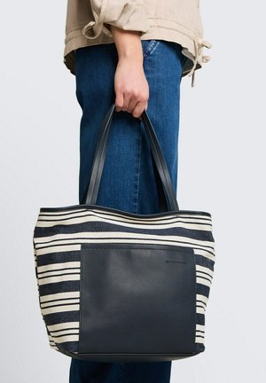 Main tenant un sac cabas rayé noir et blanc avec une poche avant noire, portant un jean bleu et une veste beige sur fond blanc.