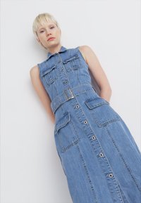 Abito di denim senza maniche, con colletto, bottoni frontali e due tasche sul petto. Colore azzurro chiaro con cinturino in vita.