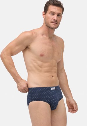 Hombre sin camiseta ajustándose unos bóxers azul marino con un pequeño patrón de rombos blancos, de pie contra un fondo claro y sencillo.