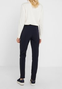 Pantalons navy ajustés avec une coupe slim, présentant une taille mi-haute et une texture lisse. Portés avec un pull de couleur claire et des baskets.