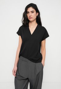 Haut noir à manches courtes avec un design en V. Tissu doux avec une texture lisse. Associé à un pantalon gris ample avec des plis.