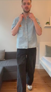 Camisa de ganga azul-claro de manga curta com botões e bolso de pala, combinada com calças largas pretas e ténis cinzentos. Fundo de pavimento em madeira.