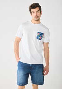 White cotton t-shirt with a navy floral pocket. Worn with denim shorts, showcasing a relaxed fit and short sleeves.