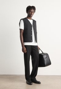 Vest matlasat negru peste un tricou alb, pantaloni negri și pantofi negri. Ținând o geantă din piele neagră. Fundal neutru.