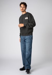 Schwarzer Sweatshirt mit "UNWIND"-Schriftzug auf der Vorderseite, kombiniert mit blauen Jeans und schwarzen Schuhen, vor einem neutralen Hintergrund.