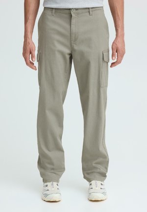 Mann trägt hellgraue Cargohosen und weiße Turnschuhe, steht mit entspannten Armen an den Seiten vor einem einfarbigen Hintergrund.