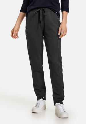 Pantalons de jogging noirs en matériau lisse et extensible. Dotés de poches zippées et d'une taille avec cordon de serrage. Associés à des baskets blanches.