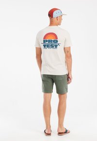 Beige t-shirt met een grafische print van een regenboogboog en vetgedrukte tekst. Groene shorts, sandalen en een meerkleurige pet maken de outfit compleet.