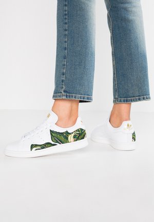 Person iført korte blå jeans og hvide sneakers med grønt bladmønster og gyldt logo, stående på hvid gulv.