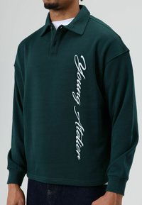 Donkergroen poloshirt met lange mouwen gemaakt van zachte stof, met een omgevouwen kraag en verticale witte geborduurde tekst aan de voorkant.