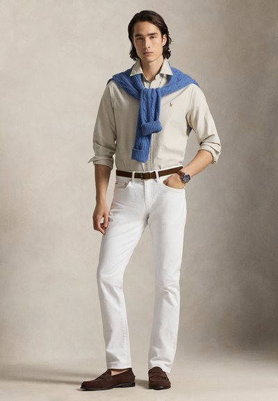 Jongetje staat met een beige overhemd met knopen, witte broek, blauwe trui over de schouders gedrapeerd, bruine riem, horloge en bruine loafers tegen een neutrale achtergrond.