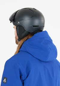 Sort skihjelm med ventiler, justerbart hagerem, båret med en blå hættejakke. Jakken har en glat tekstur og et logo på ærmet.