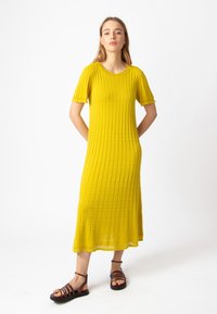 Keltainen ribbineulemekko, jossa on lyhyet hihat, puolipitkä pituus, pyöreä pääntie ja hienovarainen avoin neulontayksityiskohta. Yhdistetty ruskeisiin narusaappaisiin.