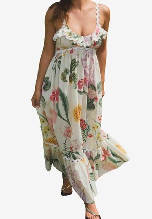 Frau trägt ein ärmelloses weißes Maxikleid mit floralem Muster und Rüschen, läuft und hält den Saum des Kleides.