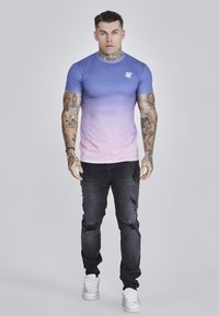 Kék és rózsaszín ombre t-shirt, rövid ujjú, testhezálló fazon, kis logóval; elhasználódott fekete farmernadrággal és fehér sportcipővel párosítva.