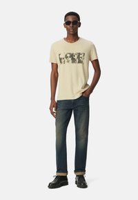 Camiseta de algodón beige con un diseño gráfico negro, combinada con unos vaqueros de mezclilla azul oscuro y zapatos negros con una suela gruesa, de pie contra un fondo blanco.