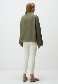 Giacca oversize verde oliva con colletto, maniche lunghe e polsini con bottoni, abbinata a pantaloni attillati bianchi e scarpe beige slip-on.