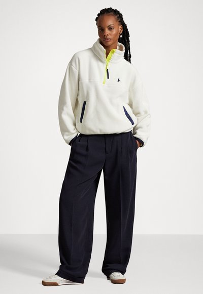 Fleece-Pullover in Off-White mit hohem Kragen, neongelbem Reißverschluss und navyblauen Akzenten; kombiniert mit lockeren navyblauen Hosen und weißen Sneakers.
