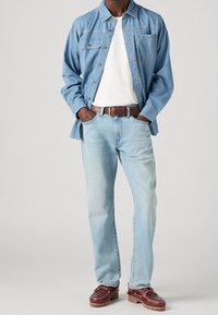 Ljusblå denimjeans och en blårandig skjorta med knappar över en vit t-shirt, kombinerat med bruna läderskor och ett brunt bälte.